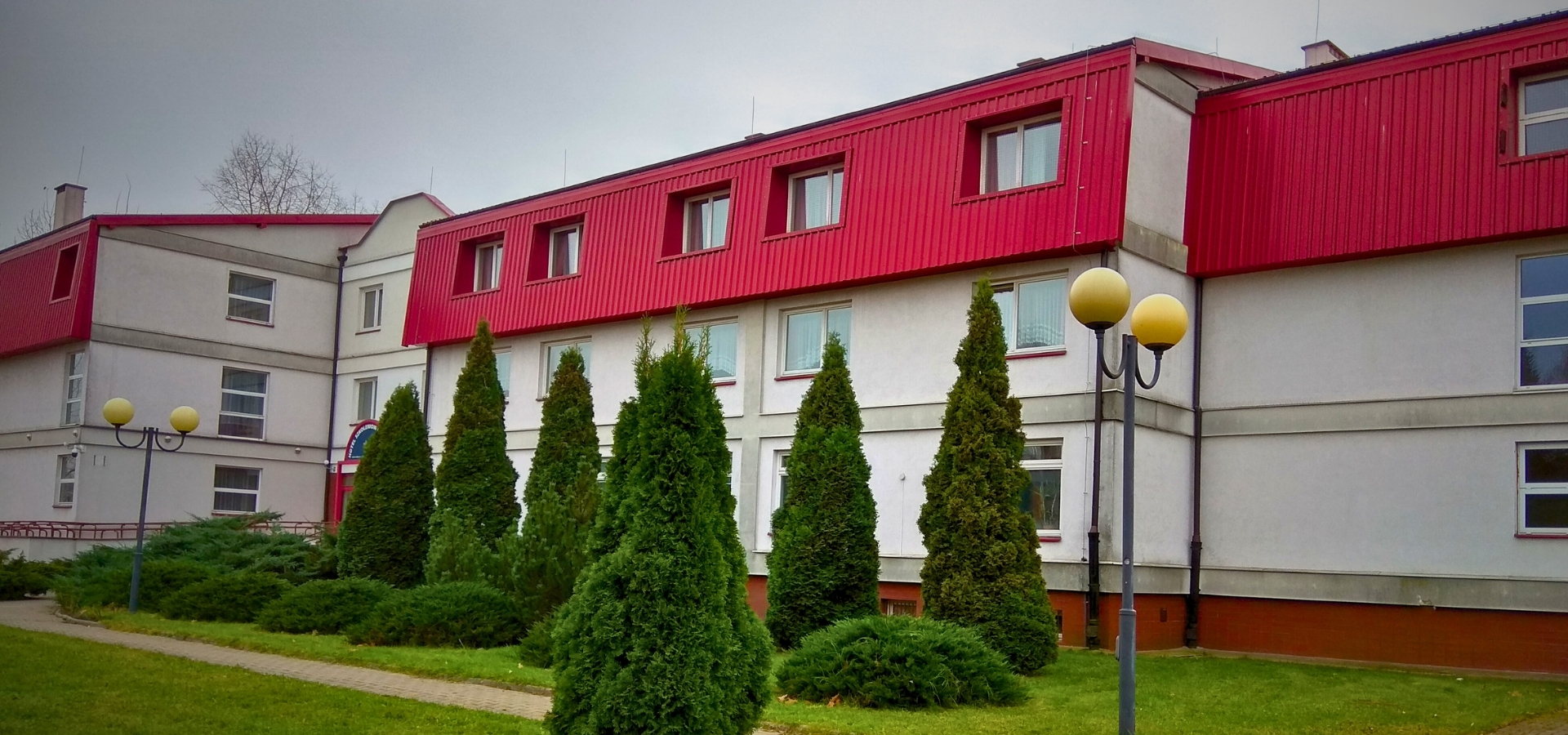 Budynek z czerwonym dachem, białymi ścianami, otoczony zielonymi drzewami i latarniami.
