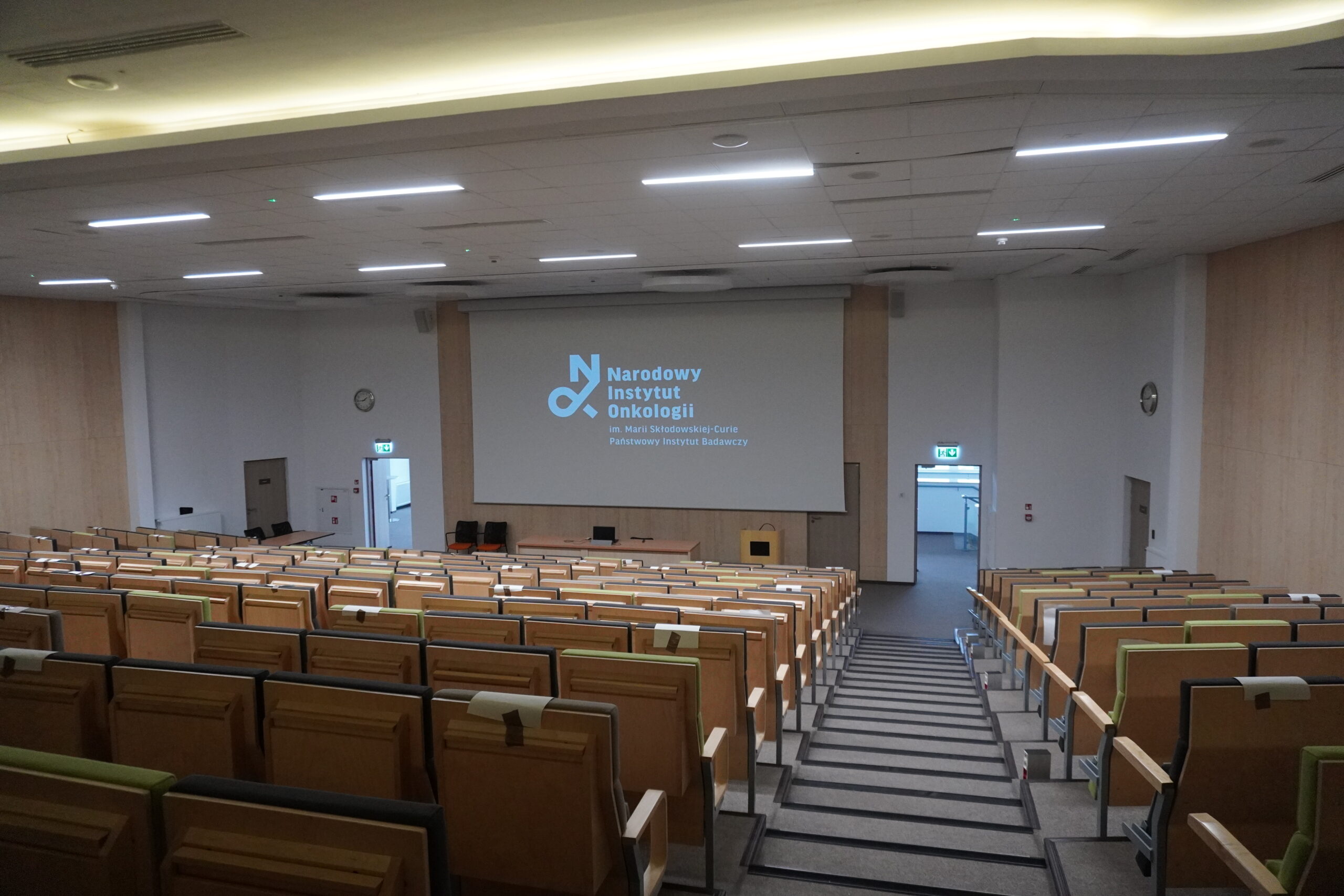 Duża sala konferencyjna z drewnianymi krzesłami, ekranem projekcyjnym i napisem "Narodowy Instytut Onkologii".