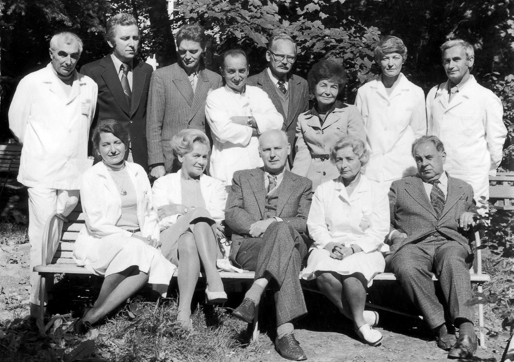 Grupa osób w formalnych strojach, część w kitlach, siedząca i stojąca na zewnątrz.