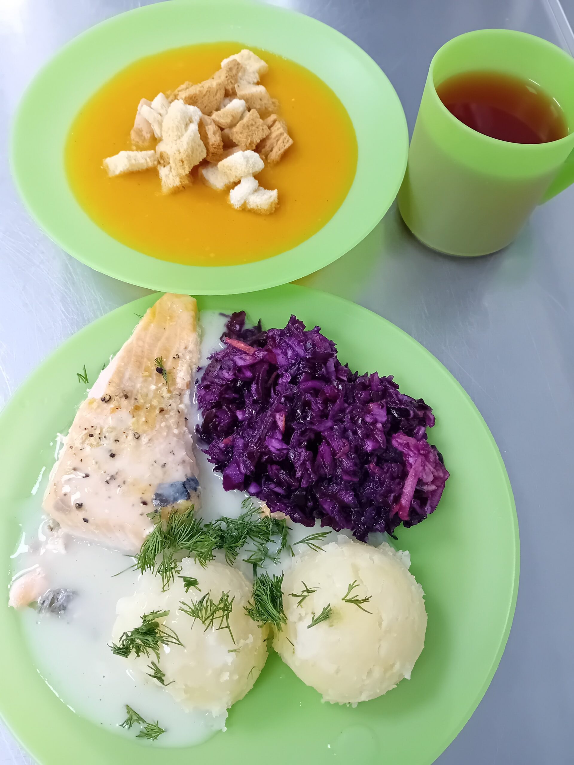 Zupa z grzankami, herbata w zielonym kubku, ziemniaki z koperkiem, ryba w sosie, czerwona kapusta na zielonych talerzach.