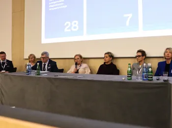 Siedem osób siedzi przy stole na tle ekranu z liczbami 28 i 7, na stole stoją butelki z wodą.