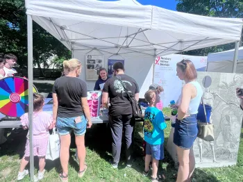 Grupa ludzi przy stoisku pod białym namiotem na pikniku, w tle drzewa i niebo.