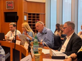 Ludzie siedzący przy stole konferencyjnym, przed nimi dokumenty i mikrofony. W tle duże okna.