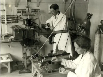 Dwie osoby w laboratorium w białych fartuchach, pracujące przy aparaturze naukowej.