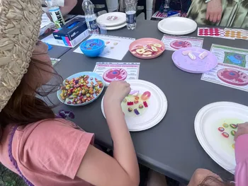 Dzieci przy stole z talerzami pełnymi kolorowych cukierków, w tle butelki z wodą i plansze.