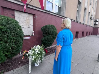 Kobieta w niebieskiej sukience stoi przed tablicą pamiątkową z kwiatami i flagami.