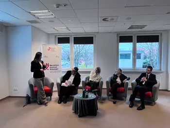 Pięć osób siedzi na krzesłach w sali konferencyjnej, jedna osoba stoi i przemawia.