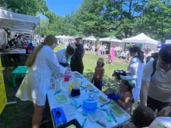 Dzieci i dorośli przy stole z eksperymentami chemicznymi na pikniku, w tle namioty i drzewa.