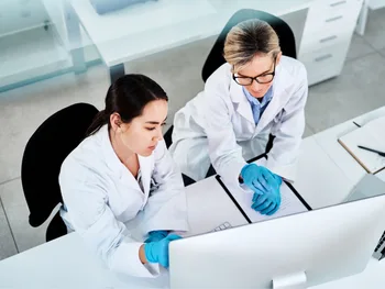 Dwie osoby w białych fartuchach i rękawiczkach przy komputerze w laboratorium.