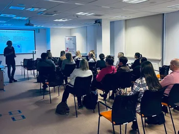 Grupa ludzi siedzi na krzesłach w sali konferencyjnej, słuchając prezentacji na ekranie.