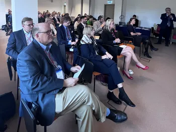 Grupa ludzi siedzących na krzesłach w sali konferencyjnej, niektórzy z identyfikatorami na szyi.