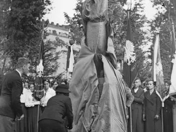 Odsłonięcie pomnika Marii Skłodowskiej-Curie przez kobietę w kapeluszu, w tle flagi i drzewa.