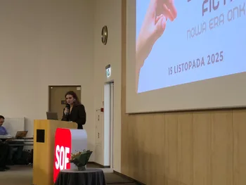 Kobieta przemawia przy mównicy na konferencji. Obok ekran z napisem: 