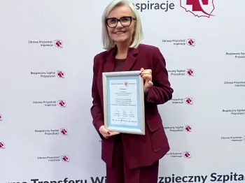 Kobieta w bordowym garniturze trzyma certyfikat na tle ścianki z logo „Zdrowa Przyszłość-Inspiracje”.