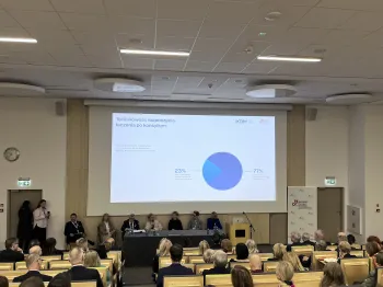 Sala konferencyjna, ludzie siedzą na krzesłach, na ekranie wykres kołowy z tekstem: „Terminowość rozpoczęcia leczenia po k…
