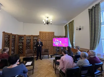 Grupa ludzi siedzi w sali z drewnianymi regałami, mężczyzna stoi przed nimi, obok ekran z kolorową grafiką.