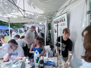 Ludzie przy stołach pod namiotem, prowadzą eksperymenty chemiczne, w tle plakat z napisem „Probówka”.