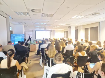 Sala konferencyjna z ludźmi siedzącymi na krzesłach, prezentacja na ekranie z przodu.