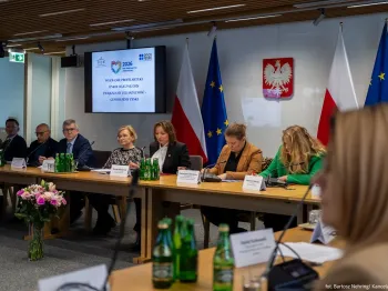 Osoby siedzą przy stole konferencyjnym, w tle flagi Polski i UE oraz ekran z tekstem.