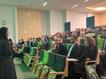 Osoby siedzące w sali konferencyjnej, jedna osoba stoi z mikrofonem, zegar na ścianie po lewej.