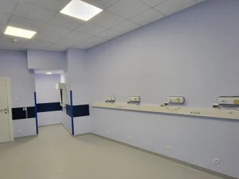 Pusta sala szpitalna z niebieskimi ścianami, białymi drzwiami i sufitowymi panelami świetlnymi.