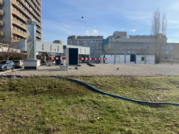 Teren z kontenerami i budynkami, w tle wysoki budynek, na pierwszym planie trawa i żwir.