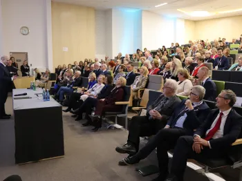 Sala konferencyjna z publicznością, osoba przemawiająca przy stole, ludzie siedzą na krzesłach w rzędach.