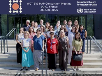 Grupa ludzi na schodach, w tle budynek. Na górze napis: „WCT EVI MAP Consortium Meeting IARC, France 26 June 2026”.
