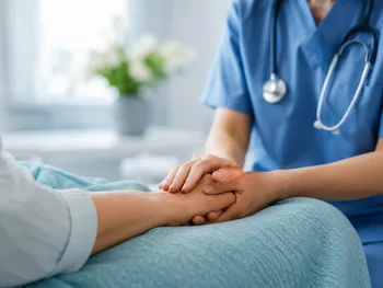 Osoba w niebieskim stroju medycznym trzyma za rękę pacjenta w łóżku szpitalnym.