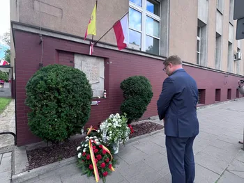Mężczyzna w garniturze stoi przed tablicą pamiątkową z wieńcami i flagami.