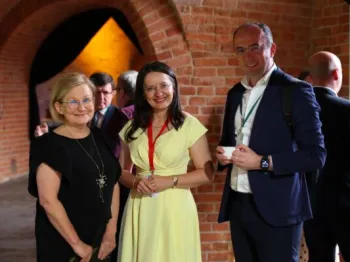 Grupa osób w eleganckich strojach stoi w ceglanej sali, uśmiechają się do kamery.