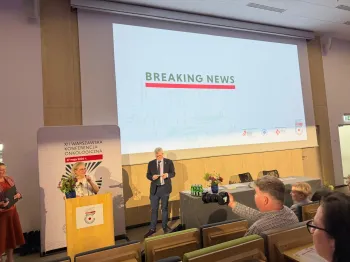 Sala konferencyjna, mówca przy mównicy, ekran z napisem 