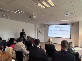 Osoba prezentuje slajd w sali konferencyjnej; widoczni uczestnicy siedzą na krzesłach.