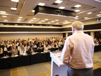 Mężczyzna przemawia do dużej grupy ludzi siedzących w sali konferencyjnej.