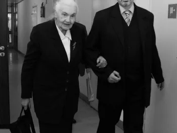 Starsza kobieta i mężczyzna w garniturze idą korytarzem, trzymając się pod ramię.