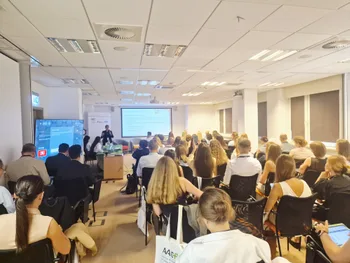 Sala konferencyjna z uczestnikami siedzącymi na krzesłach, prezentacja na ekranie z przodu.