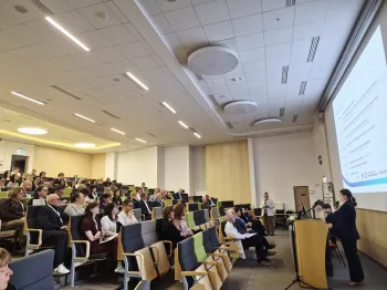 Sala konferencyjna z osobami siedzącymi na krzesłach, osoba przemawia przy mównicy, ekran z prezentacją.