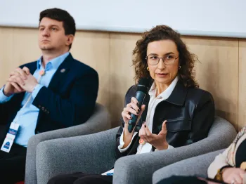Kobieta z mikrofonem siedzi na szarym fotelu, obok niej mężczyzna w garniturze.