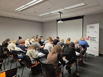 Sala szkoleniowa z osobami piszącymi notatki, w tle ekran i roll-up Narodowego Instytutu Onkologii