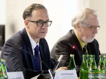 Dwaj mężczyźni w garniturach siedzą przy stole z mikrofonami i butelkami wody.