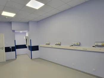 Pusta sala szpitalna z niebieskimi ścianami, panelami z gniazdkami i lampami na ścianie.