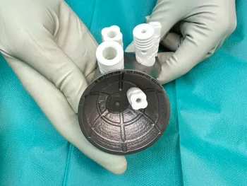 Ręce w rękawiczkach trzymają okrągły, plastikowy element z kilkoma białymi rurkami.