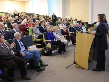 Osoba przemawia przy mównicy do zgromadzonej publiczności w sali konferencyjnej.