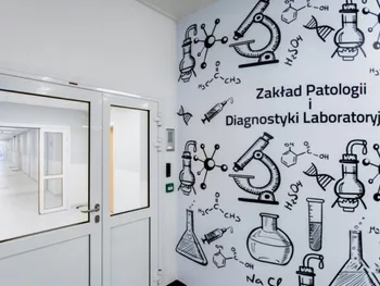 Korytarz z białymi drzwiami, na ścianie napisy: „Zakład Patologii i Diagnostyki Laboratoryjnej” oraz rysunki sprzętu labor…