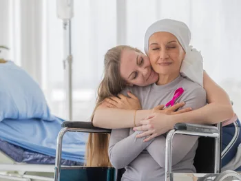 Kobieta w chuście na głowie siedzi na wózku inwalidzkim, przytula ją młodsza kobieta.