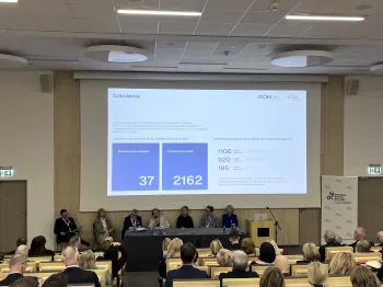 Panel dyskusyjny w sali konferencyjnej, ekran z prezentacją o szkoleniach, siedem osób przy stole.