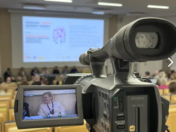 Kamera nagrywająca konferencję w sali wykładowej, na ekranie prezentacja i panel dyskusyjny.
