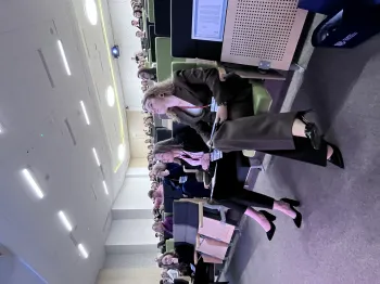 Osoby siedzące w audytorium, patrzące w stronę sceny lub ekranu, z notatkami na kolanach.