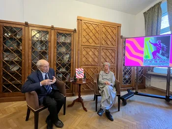 Dwie osoby siedzące na krzesłach w bibliotece, obok ekran z kolorową grafiką i książka na stoliku.