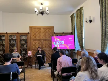 Grupa ludzi siedzi w sali z drewnianymi regałami, dwie osoby prowadzą prezentację przy ekranie.
